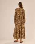 A woman with long brown hair wears the natalie martin Fiore Maxi, an ankle-length, long sleeve A-line dress in tan and black leopard print, paired with flat sandals. She stands facing away against a neutral background.