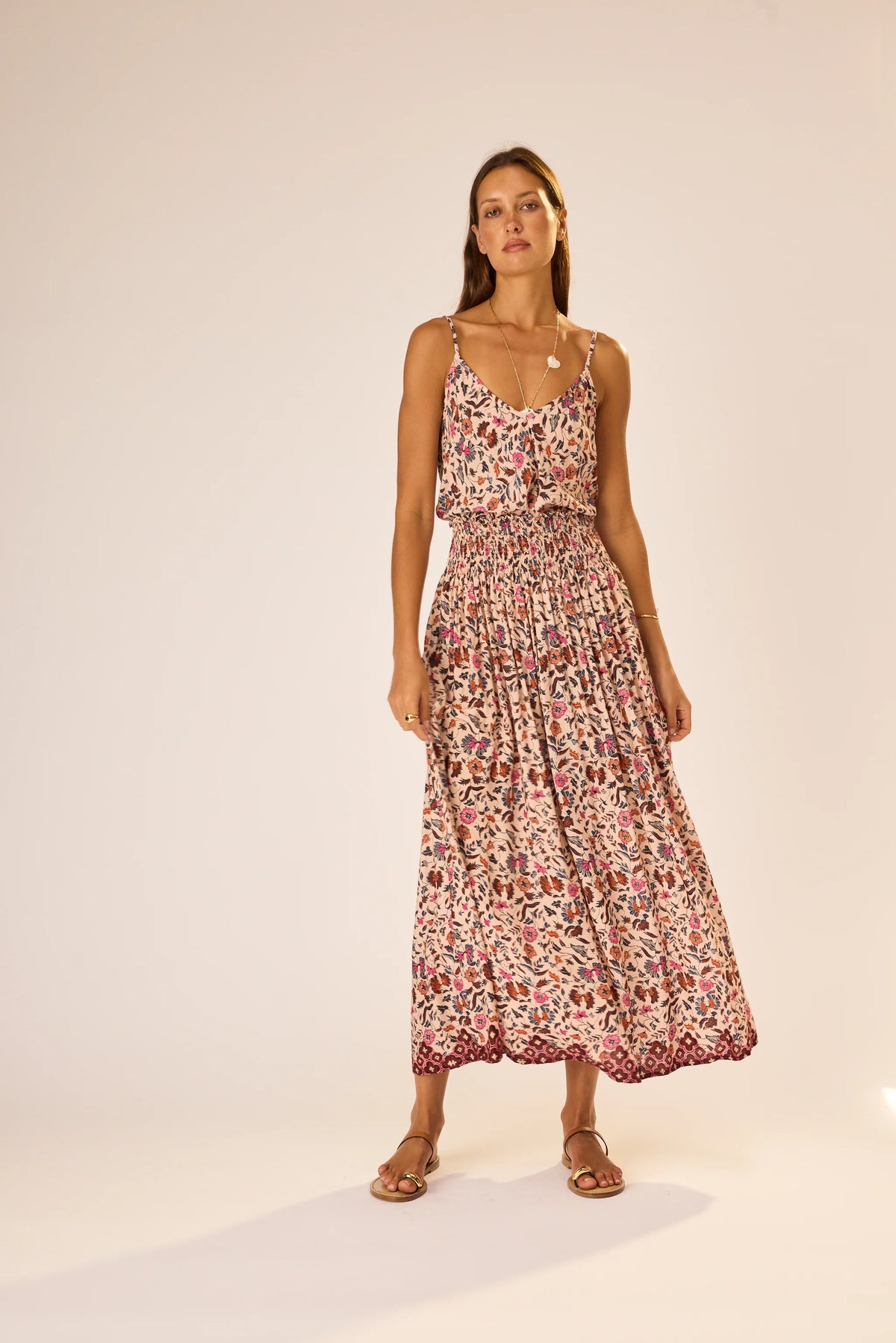 A woman wears the natalie martin Bella Skirt, a colorful floral rayon piece with an elastic waist, styled with flat sandals and layered necklaces. She has long hair and stands against a softly lit plain background.
