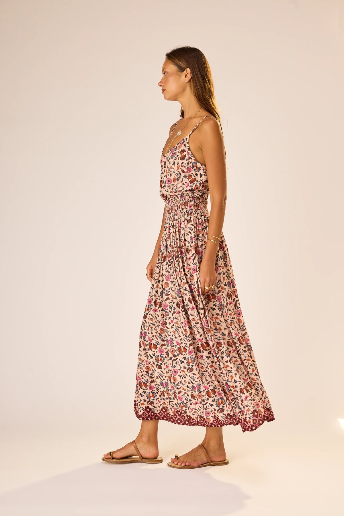 A woman stands in profile wearing the natalie martin Bella Skirt, an ankle-length floral rayon skirt with an elastic waist, paired with flat sandals. She has long hair and poses against a plain, light-colored background.