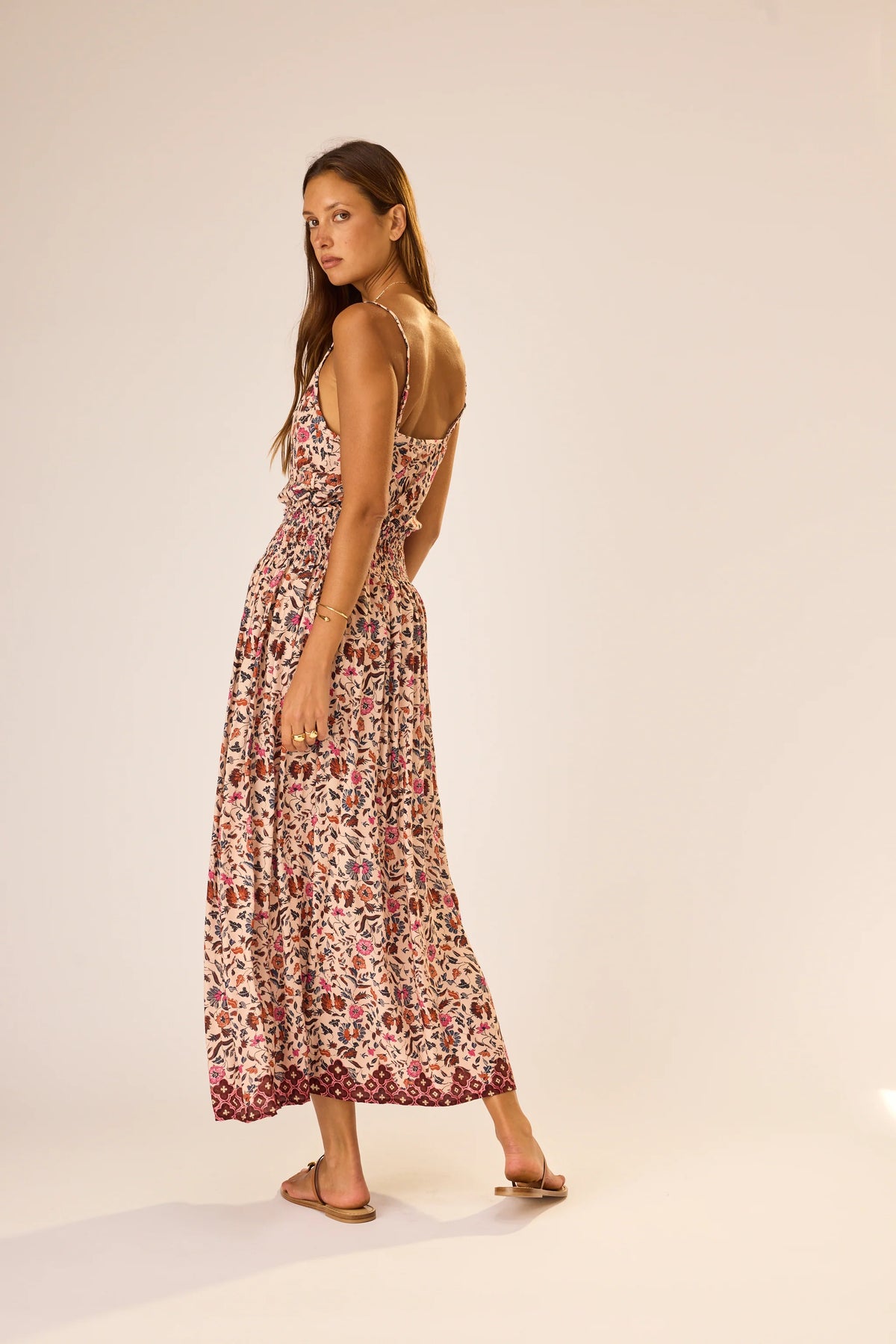 A woman wears the natalie martin Bella Skirt, a long floral-patterned rayon skirt with an elastic waist, paired with sandals. She stands on a plain background, looking back over her shoulder with a neutral expression.