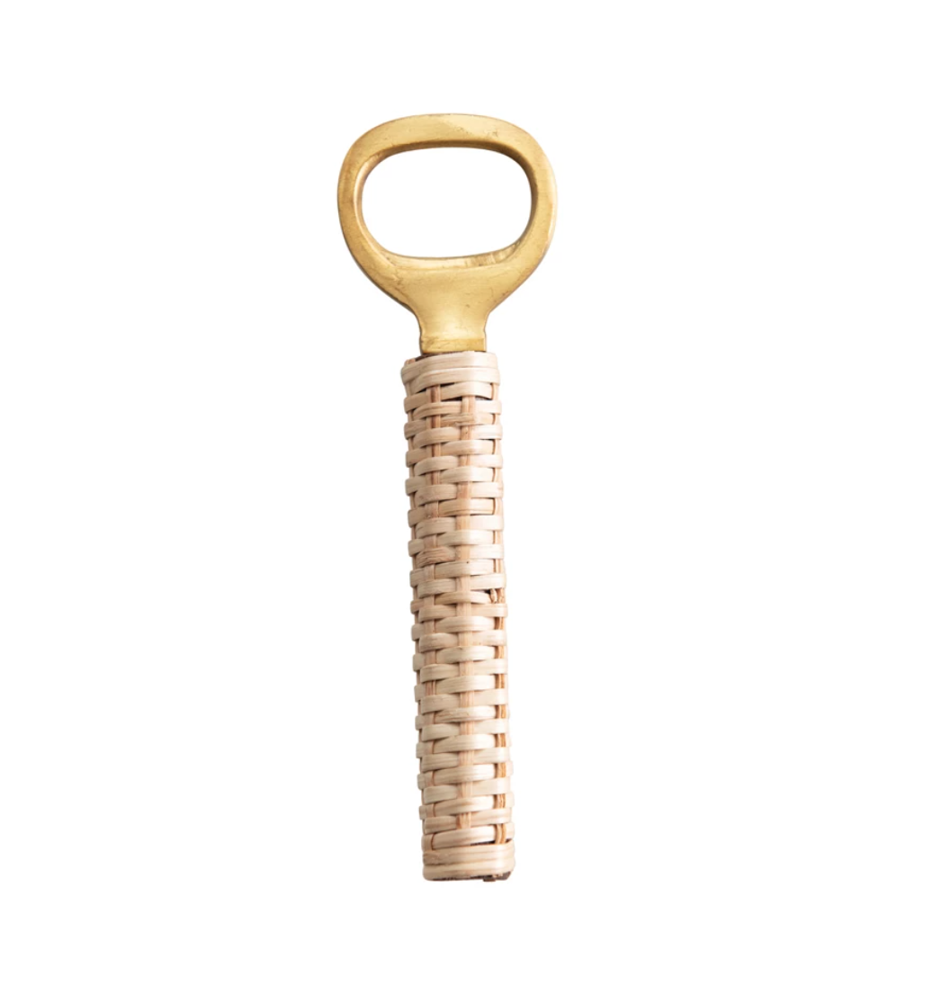The Creative Co-op Bottle Opener features a brass design with an oval-shaped opening for bottle caps and a woven rattan handle, adding a rustic touch. Its light brown, cylindrical grip ensures comfort during use. Perfect as a stocking stuffer gift, it is pictured against a white background.