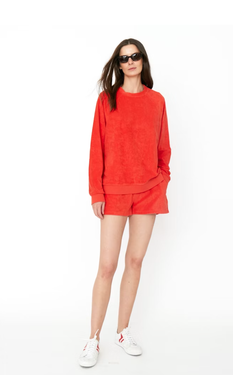 A woman in a vibrant red sweater and Kule's Terry Venus shorts set, wearing white sneakers and sunglasses, stands confidently against a white background.