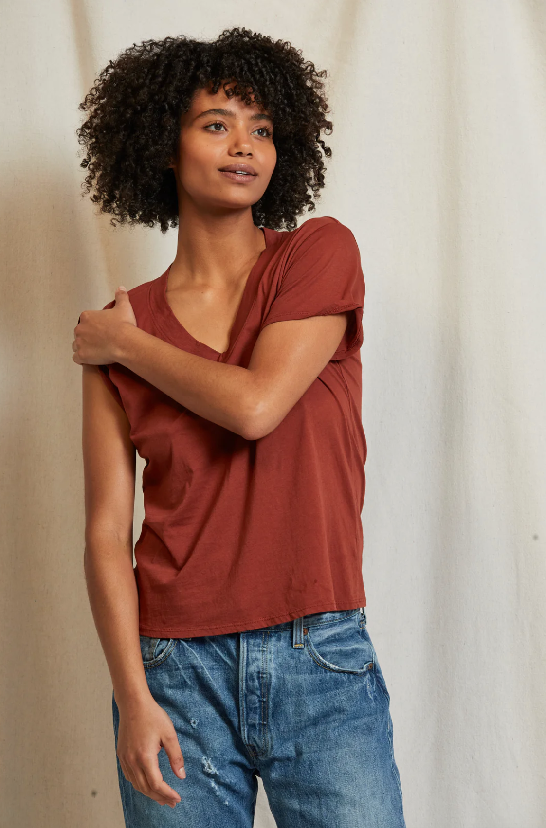 A person with curly hair is standing against a plain background. They are wearing a Hendrix Boxy V-Neck Tee by Perfectwhitetee in a reddish-brown shade, with rolled-up sleeves, paired with blue jeans. The 100% cotton shirt is a staple piece in their wardrobe. The person has one arm crossed over to touch the opposite shoulder and they are looking slightly to the side with a relaxed expression.