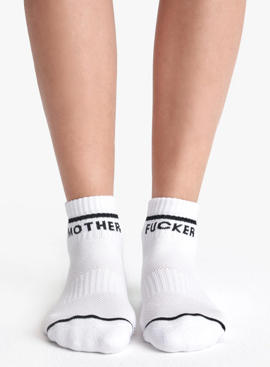 Close-up of a person's lower legs and feet wearing black and white Baby Steps Ankle socks. Each sock has a black stripe near the top and text; the left sock reads "MOTHER" and the right sock reads "FUCKER.