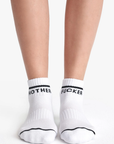 Close-up of a person's lower legs and feet wearing black and white Baby Steps Ankle socks. Each sock has a black stripe near the top and text; the left sock reads "MOTHER" and the right sock reads "FUCKER.
