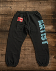 The MadeWorn P.E. Sweatpants by Made Worn are unisex black sweatpants made from heavyweight French terry cotton. They feature "ARTIST" in bold blue letters on the right leg and a small red "SPRAYED GOOD HANDWORK" patch on the left hip. Designed in vintage style, they are crafted in Los Angeles.