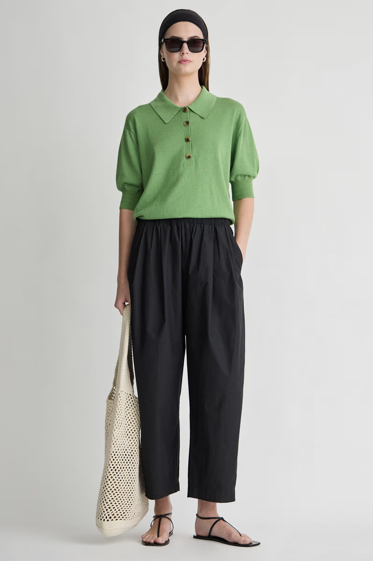 A person wearing a green collared knit top, Apiece Apart Spa Pleat Pant in black, black sandals, sunglasses, a black headband, and holding a cream-colored net tote bag stands with hands in pockets.