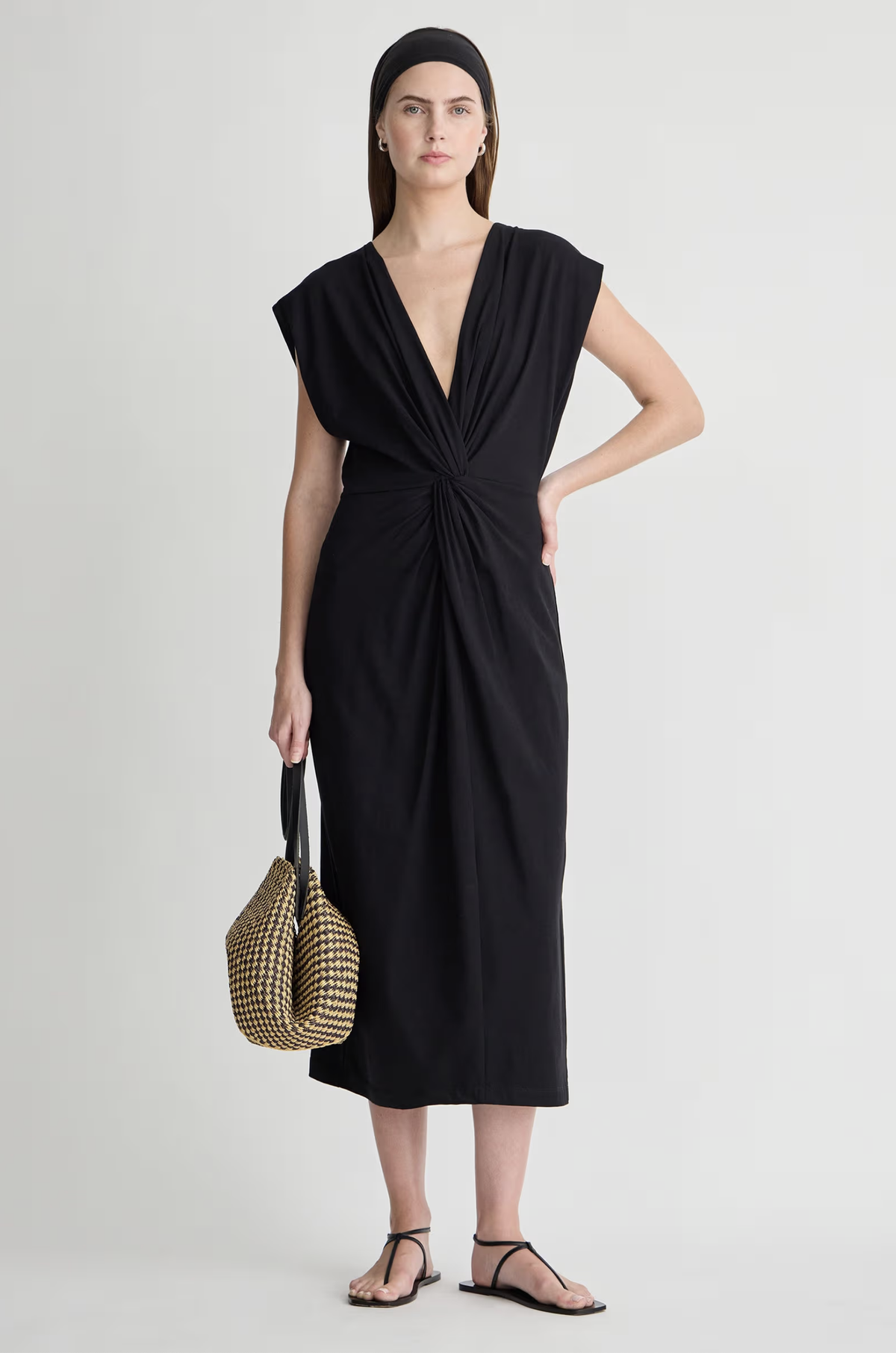 A woman wears the Apiece Apart Margit Twist Dress, featuring a sleeveless deep V-neck with front twist detail, paired with black sandals, a black headband, and a two-handle woven bag against a light background.