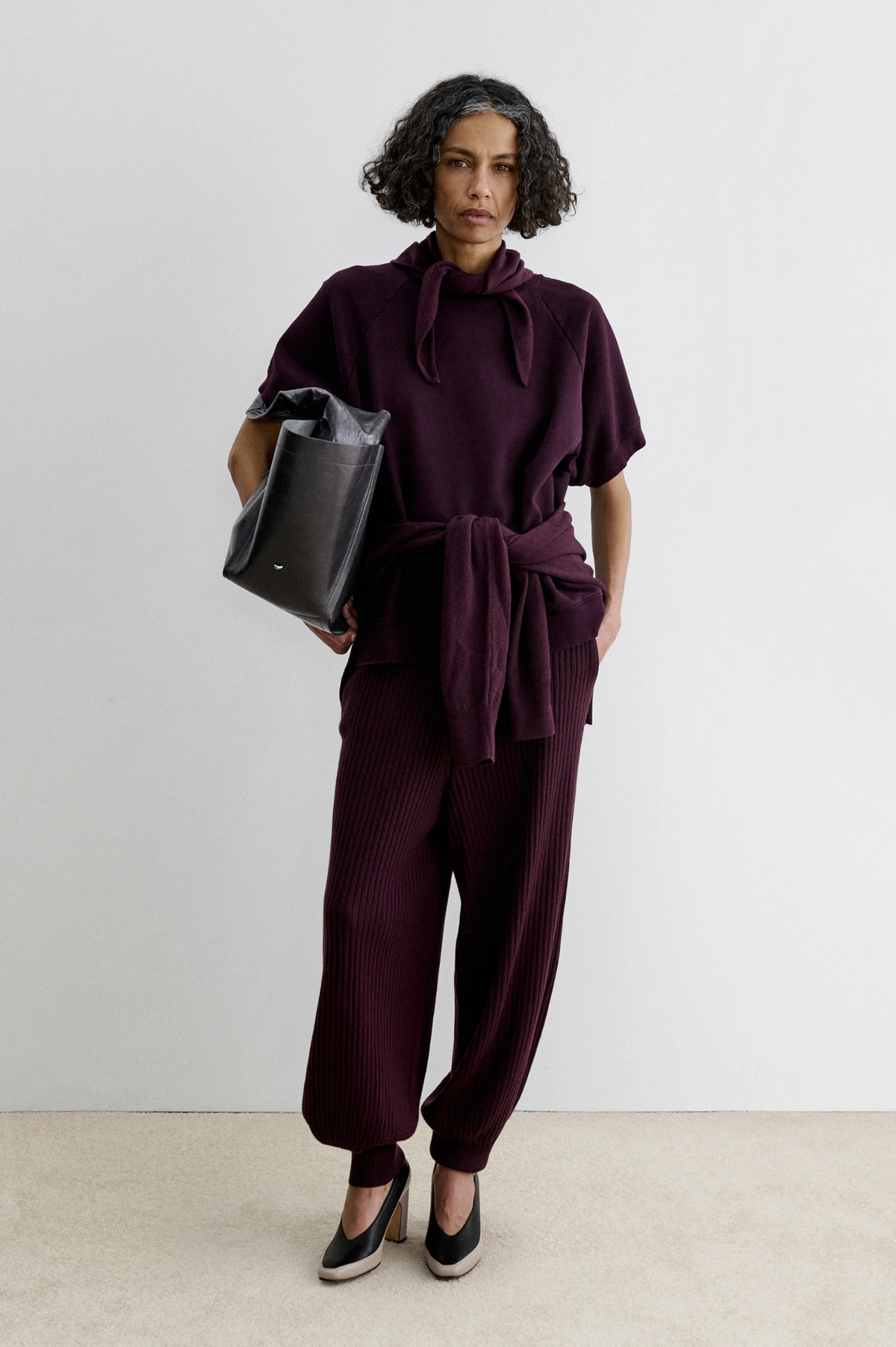Rachel Comey Nim Pant in burgundy, high-waisted and relaxed fit, styled with a black bag against a white wall.