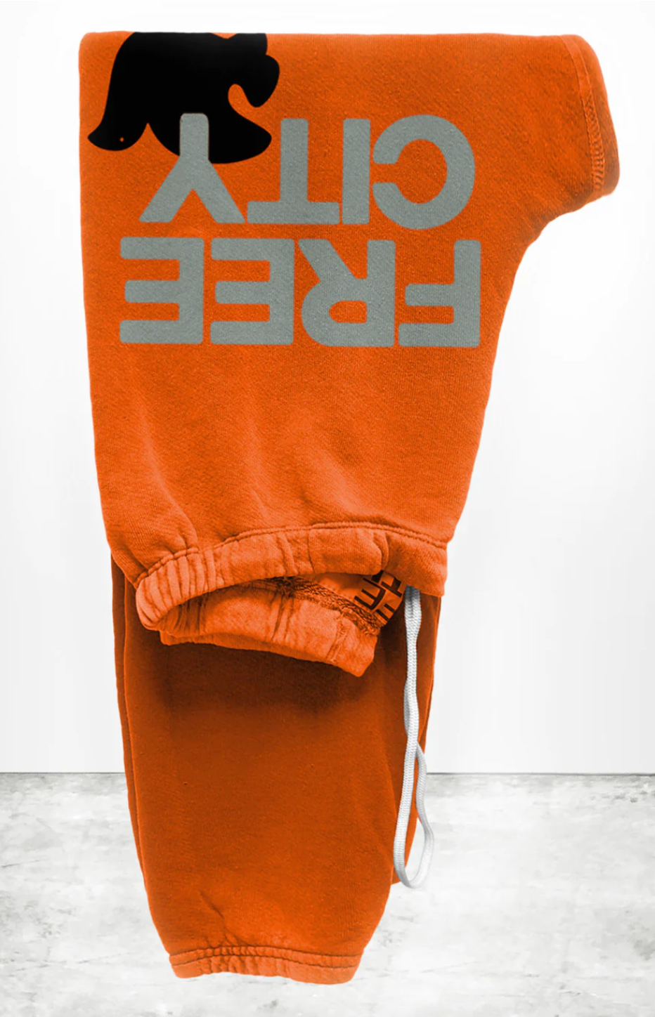 An orange pair of Free City (sparrow, LLC) FREECITY Large Sweatpant made from lightweight French terry, featuring the text "FREE CITY" in large, grey letters and a black silhouette of a small animal on the side. The unisex FLOW sweatpant is folded neatly, showcasing the text and design prominently against a plain grey and white background.