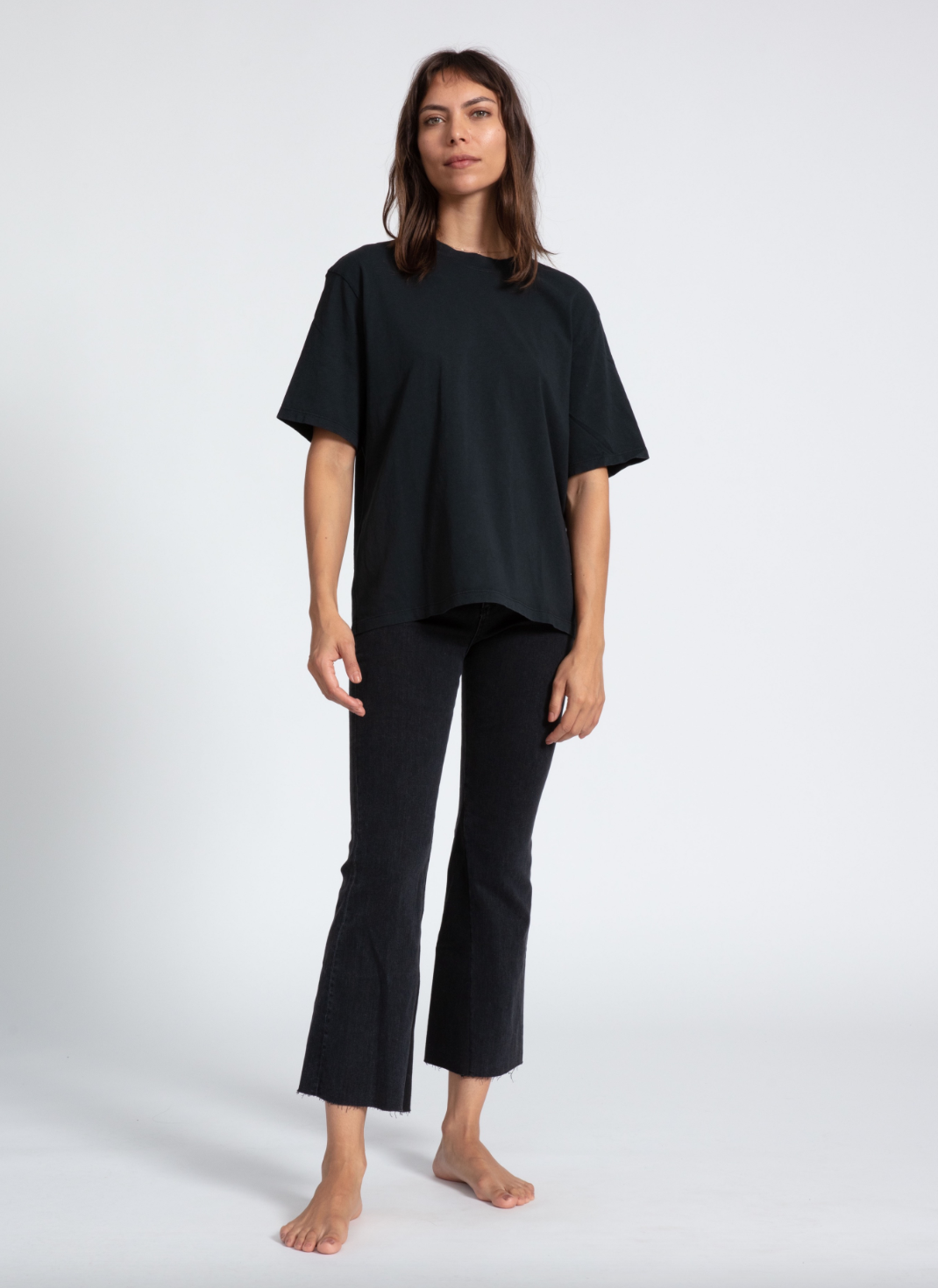 A woman stands against a plain background in a Scottsdale Arizona bungalow, wearing a loose-fitting black ASKK boy tee and dark cropped jeans, barefoot, looking directly at the camera with a subtle expression.