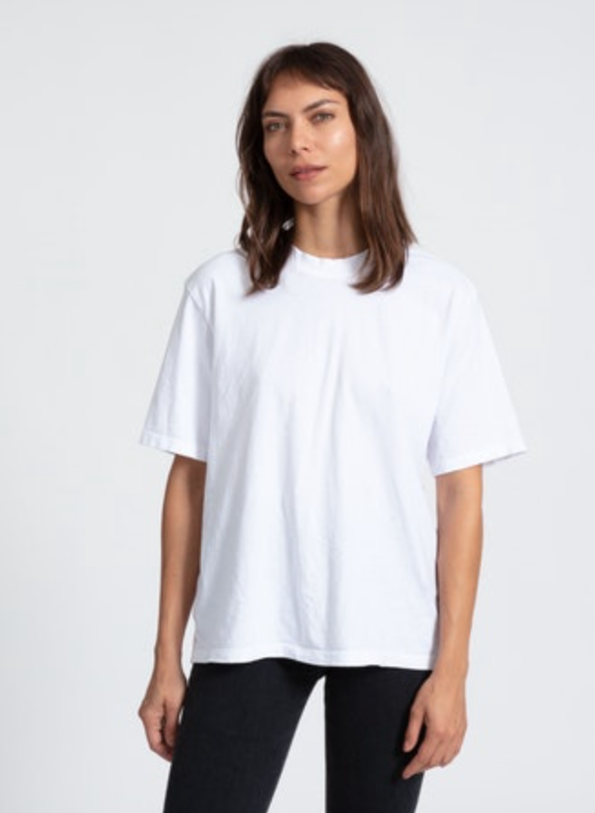 A woman with shoulder-length brown hair stands wearing a plain white ASKK boy tee and black pants against a light gray background in Scottsdale, Arizona. She looks directly at the camera with a neutral expression.