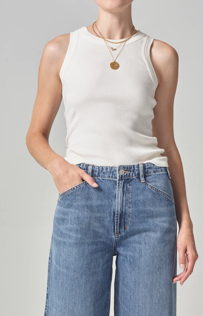 A woman in a sleeveless white ribbed racerback Isabel Rib Tank by Citizens Of Humanity/AGOLDE and blue jeans, with one hand casually tucked into the pocket. She wears a simple golden necklace. The background is plain and light-colored.