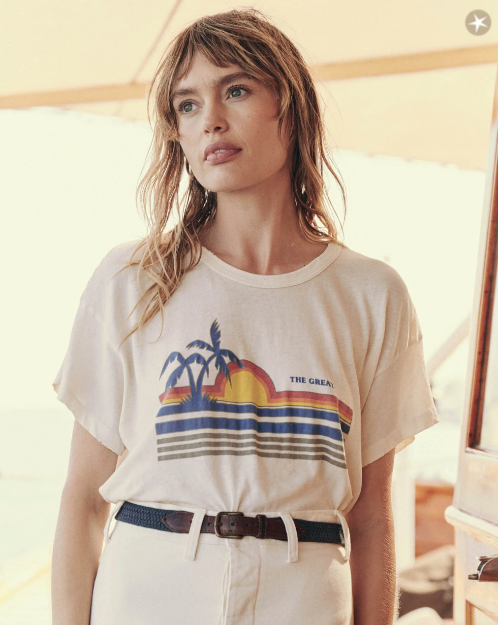 A woman with long, wavy hair stands indoors wearing a white vintage tee featuring a sunset, palm trees, and stripes. She pairs it with high-waisted white pants and a dark belt. With a contemplative expression, she gazes slightly off-camera, showcasing The Boxy Crew by The Great Inc.
