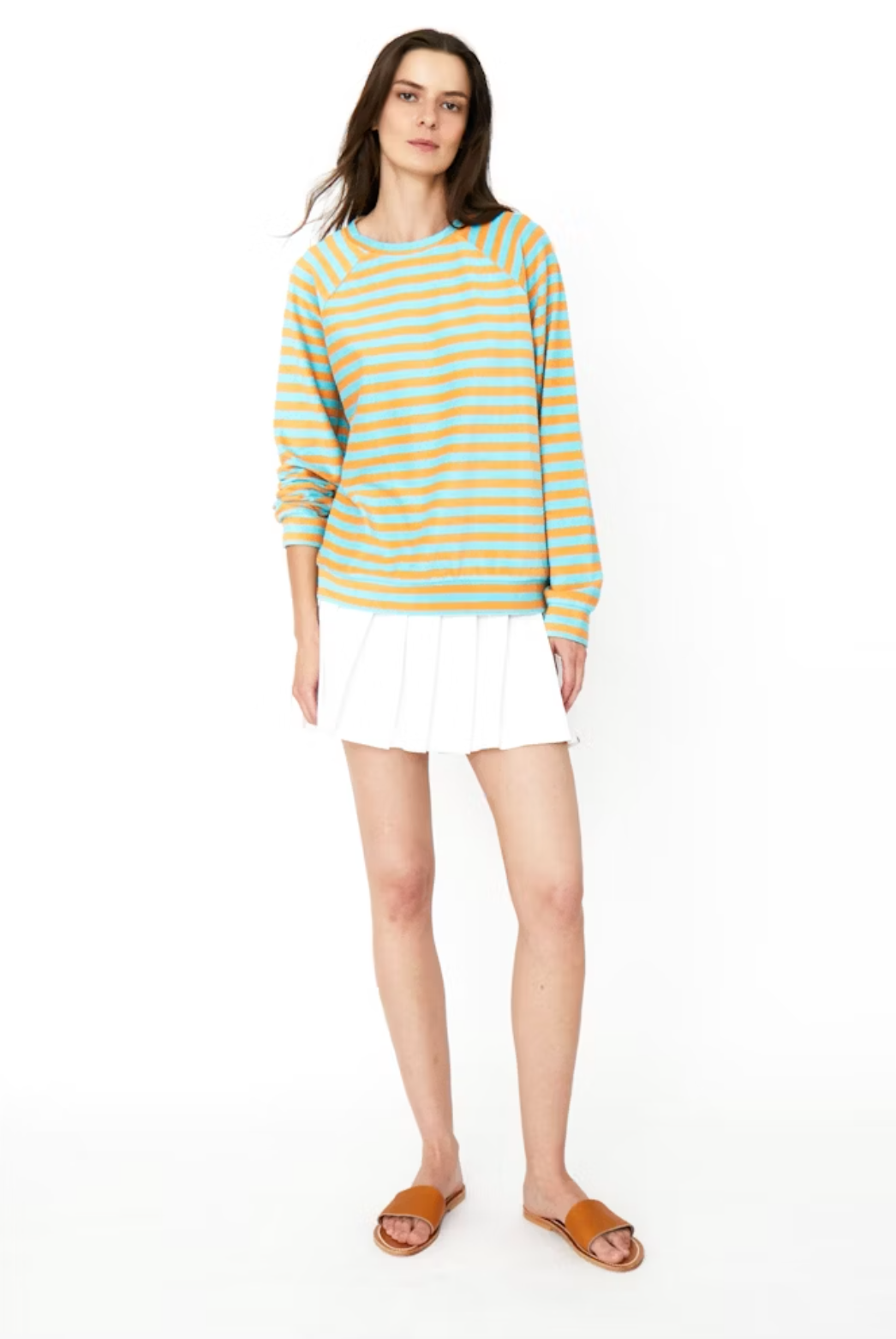 A person with long hair, wearing a blue and yellow striped The Terry Franny sweatshirt by Kule paired with a white pleated skirt and brown sandals, stands against a plain white background. They have a relaxed and casual stance.