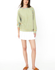 A person with long hair, wearing a blue and yellow striped The Terry Franny sweatshirt by Kule paired with a white pleated skirt and brown sandals, stands against a plain white background. They have a relaxed and casual stance.
