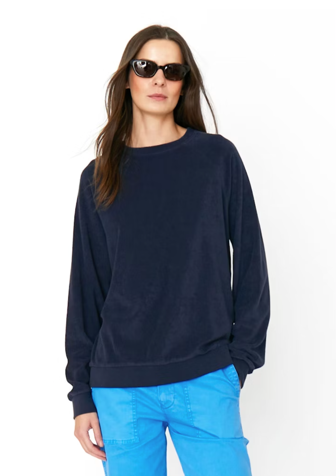 A woman with long, dark hair wearing black sunglasses and a navy blue Kule The Terry Franny sweatshirt with raglan sleeves. Her bright blue pants contrast the plain white background as she stands confidently with her hands in her pockets.