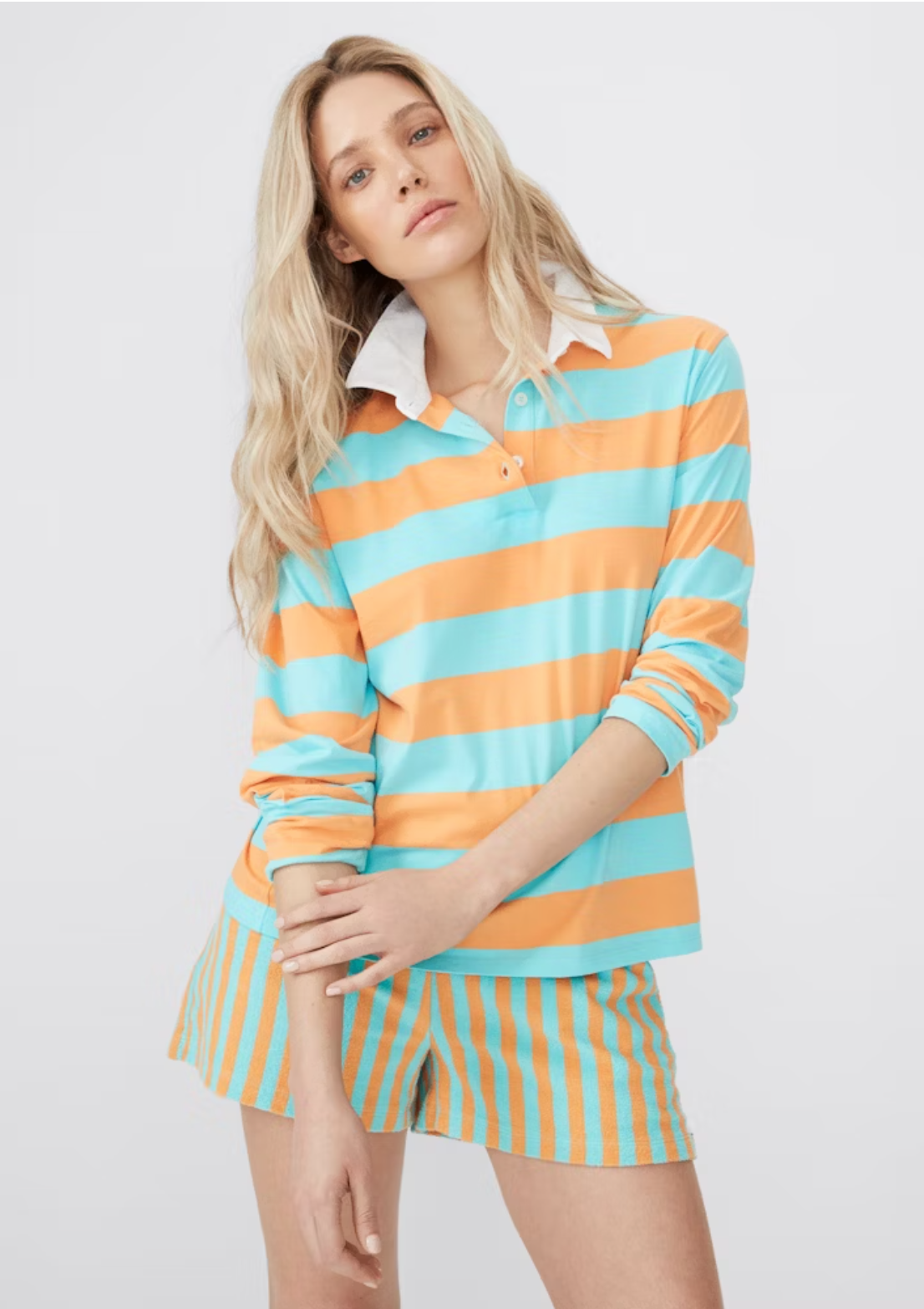A person with long blonde hair is wearing a collared cotton-blend shirt with orange and teal wide horizontal stripes. The shirt has long sleeves and is paired with matching terry shorts featuring an elastic waist and vertical stripes in the same colors, known as The Terry Venus Short by Kule. The background is plain and light-colored.