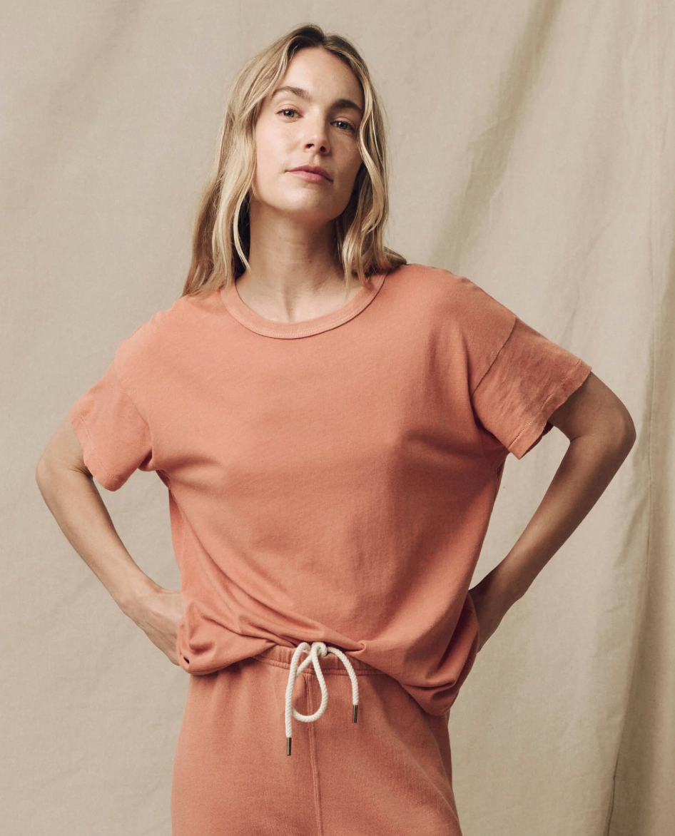 A person with long blonde hair stands confidently with hands on hips. They are wearing a matching, 100% cotton peach-colored outfit with a relaxed fit, featuring The Boxy Crew by The Great Inc., a short-sleeved top, and pants that feature a drawstring. The background is a neutral beige canvas.