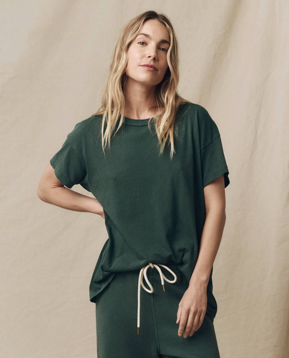 A person sports a loose-fitting, dark green tee reminiscent of a vintage style with matching drawstring pants. They have long, wavy blonde hair and stand against a beige fabric backdrop with one hand on their hip, gazing directly at the camera. The tee they are wearing is The Boxy Crew by The Great Inc.