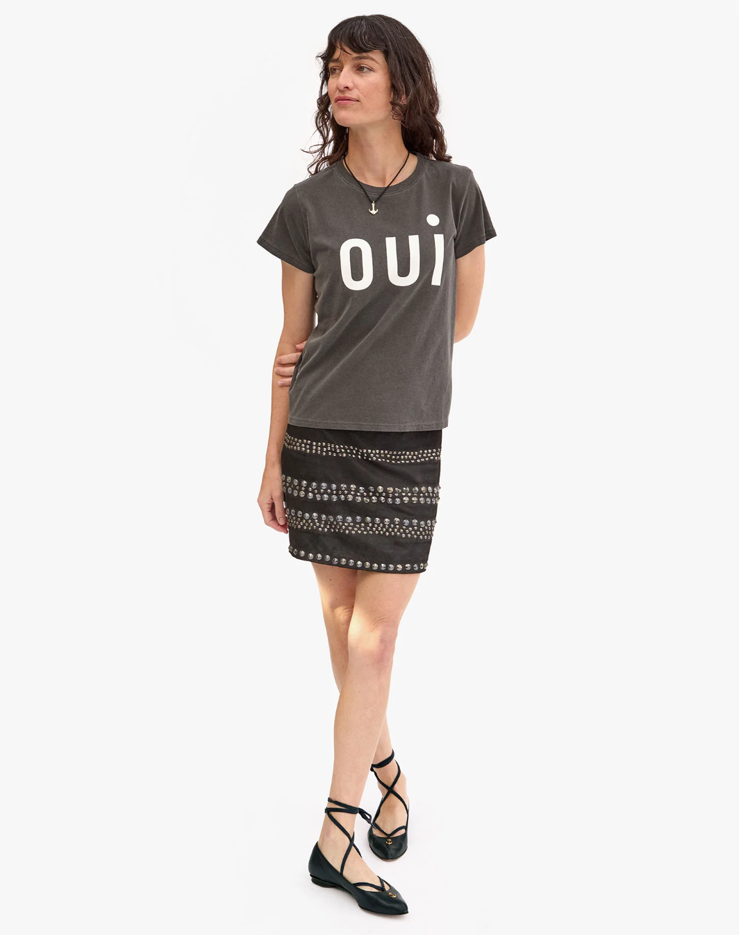 A person with long dark hair stands against a plain background. They are wearing a pre-shrunk, 100% cotton T-shirt with the word "OUI" printed on it, specifically the Classic Tee - OUI by Clare Vivier, and a patterned skirt. They have black ballet flats with ankle straps and are accessorized with a necklace and a bracelet.