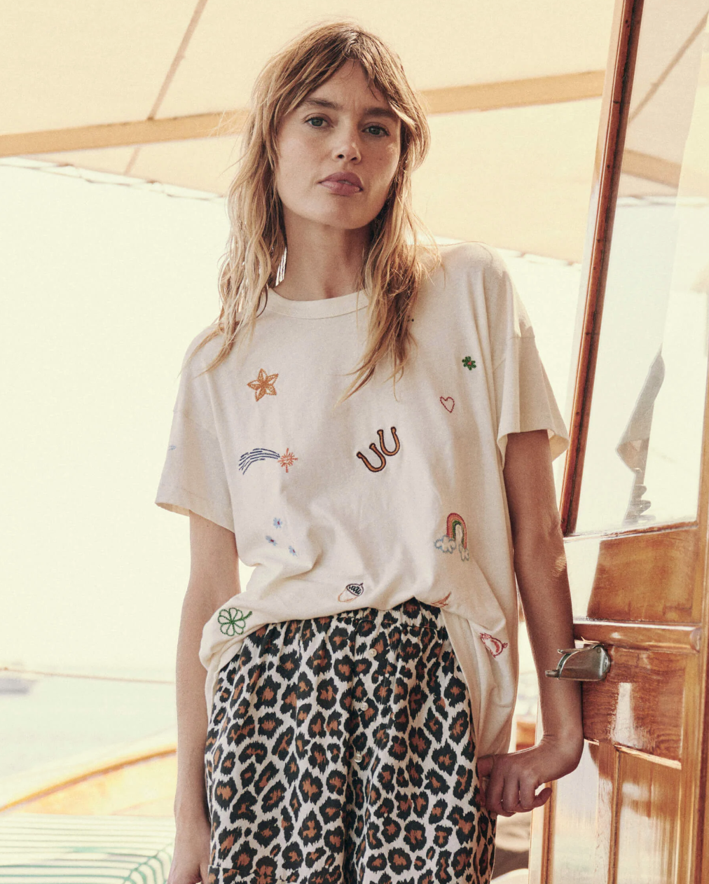 A young woman with long, wavy hair stands leaning against a wooden door on a boat. She is wearing The Boxy Crew by The Great Inc. and leopard print pants that give off vintage tees vibes. The background shows a bright, sunny day on the water.
