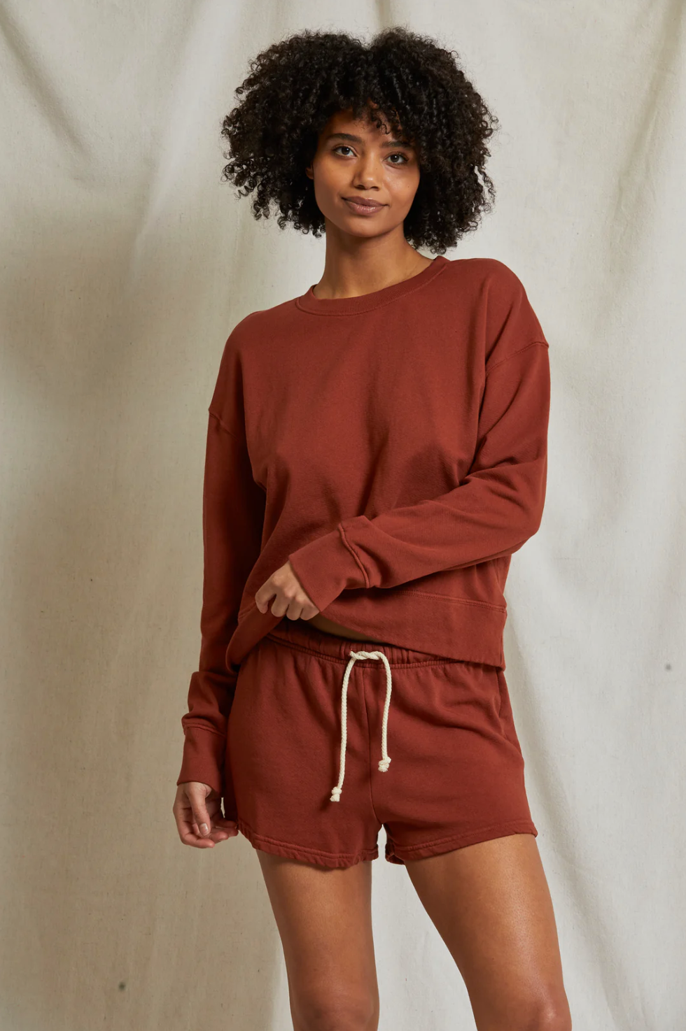 A person with curly hair stands in front of a beige background, wearing a long-sleeved, rust-colored sweatshirt and matching Perfectwhitetee Layla Terry Shorts with an elastic waistband. They are smiling slightly and looking at the camera, with one hand resting on their hip and the other on their wrist.