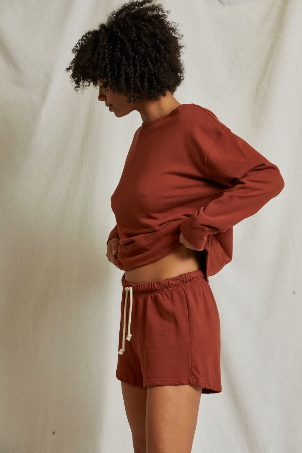 A person stands against an off-white backdrop, adjusting their maroon long-sleeve shirt. They are wearing Perfectwhitetee's Layla Terry Shorts made from French terry cotton, featuring a white drawstring and elastic waistband. With curly hair and a slightly downward gaze, they maintain a relaxed posture.
