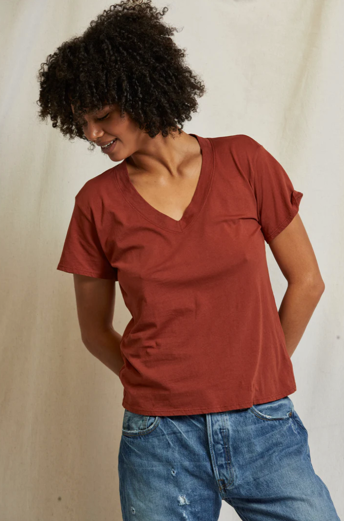 A person with curly hair is standing against a light background, looking down and to the side while smiling. They are wearing a rust-colored Hendrix Boxy V-Neck Tee from Perfectwhitetee and blue jeans, an essential staple, with hands held behind their back.