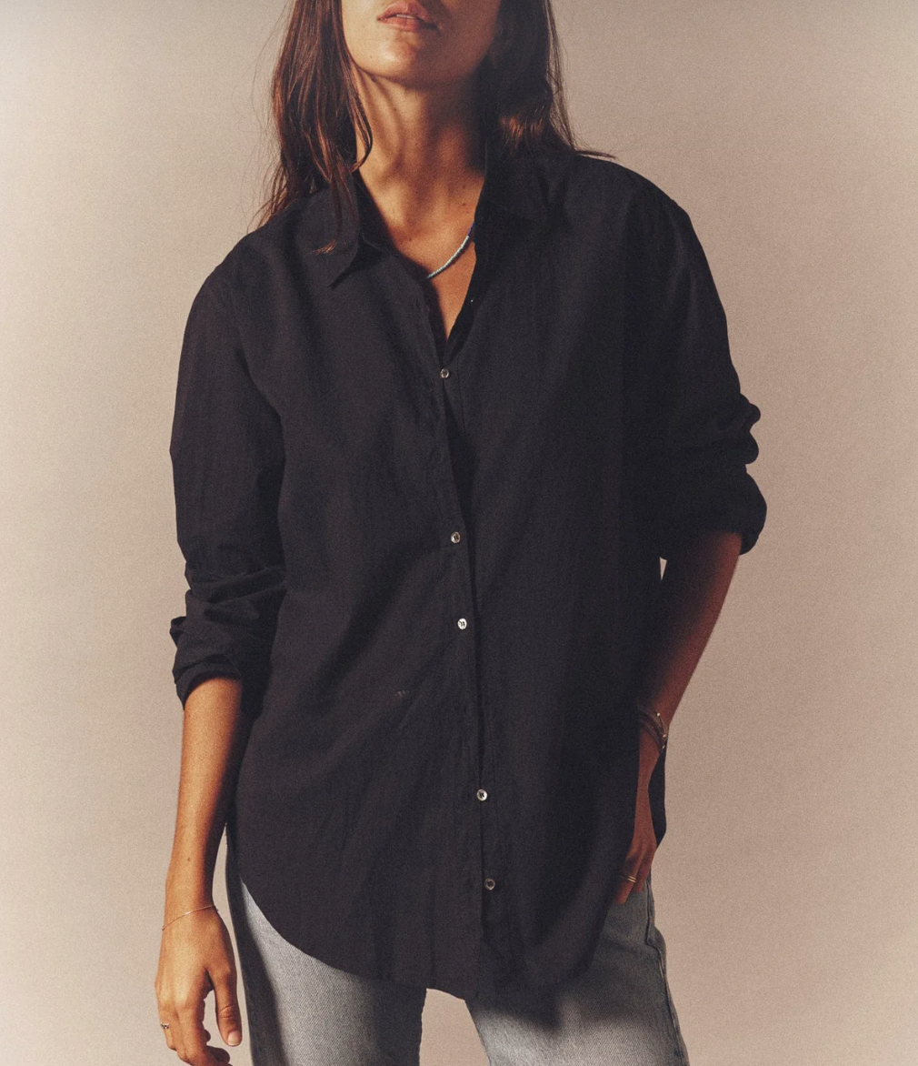A person with long hair is wearing a loose, dark Xirena Beau Shirt and light blue jeans. They are standing against a neutral background with their left hand in their pocket. The scene feels casual and relaxed, capturing the quintessential essential look effortlessly.