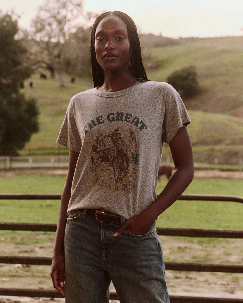 A person with long braided hair stands outdoors in front of a fence, wearing The Great Inc.'s Boxy Crew vintage tee in gray, which features a graphic of a horse and rider and the text "The Great." They have their right hand resting on their waist and are dressed in jeans with a belt. Behind them, a scenic, grassy landscape unfolds.