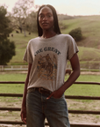 A person with long braided hair stands outdoors in front of a fence, wearing The Great Inc.'s Boxy Crew vintage tee in gray, which features a graphic of a horse and rider and the text "The Great." They have their right hand resting on their waist and are dressed in jeans with a belt. Behind them, a scenic, grassy landscape unfolds.