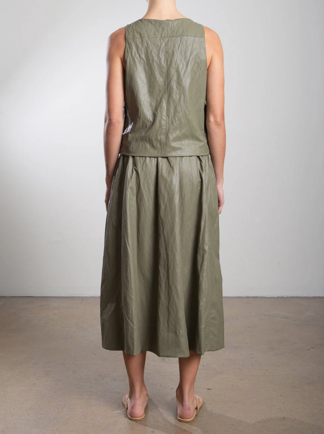 Wearing a sleeveless green dress featuring an elastic waist and gathered midi skirt, made of rough-textured fabric, a person stands with their back to the camera in a plain room. Beige sandals complete the look. The skirt is known as the Claudia Skirt by Pharaoh.