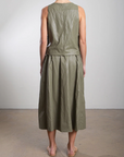 Wearing a sleeveless green dress featuring an elastic waist and gathered midi skirt, made of rough-textured fabric, a person stands with their back to the camera in a plain room. Beige sandals complete the look. The skirt is known as the Claudia Skirt by Pharaoh.