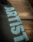 A close-up shows MadeWorn P.E. Sweatpants, a black French terry cotton fabric with "ARTIST" in large blue capital letters, offering a vintage unisex fit as it rests on a rustic wooden surface.