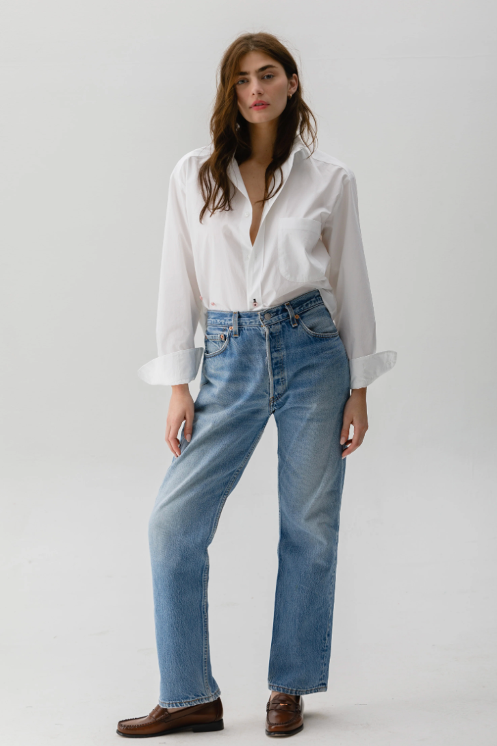 A person stands against a white background, effortlessly styled in Cissa's relaxed-fit Daily Shirt made from paper cotton with rolled-up sleeves, paired with comfy blue jeans. Their long brown hair complements the earthy tones of their brown loafers.