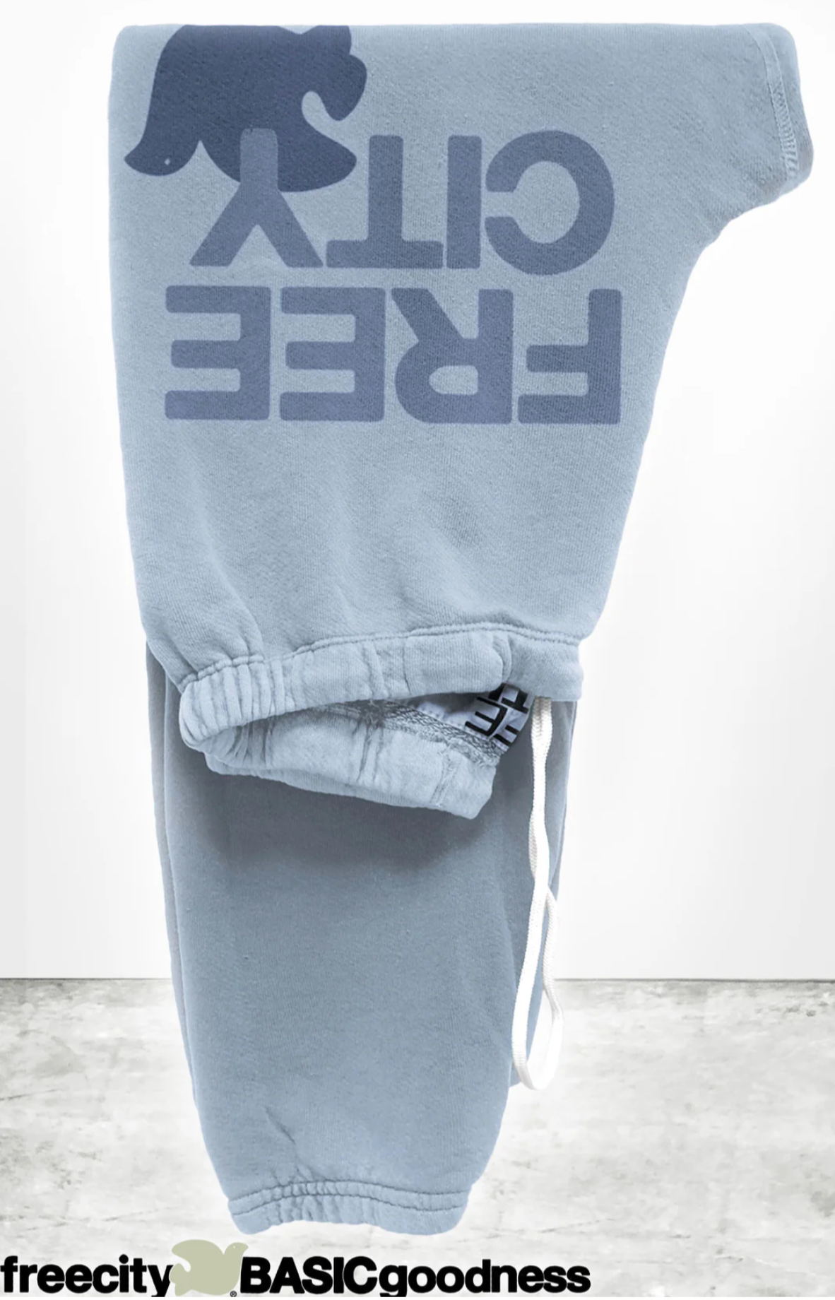 The FREECITY Large Sweatpant by Free City (sparrow, LLC) features a light blue color, dark blue "FREE CITY" print with bird graphic, unisex fit, white drawstring waistband, minimal design, and is proudly made in the USA.