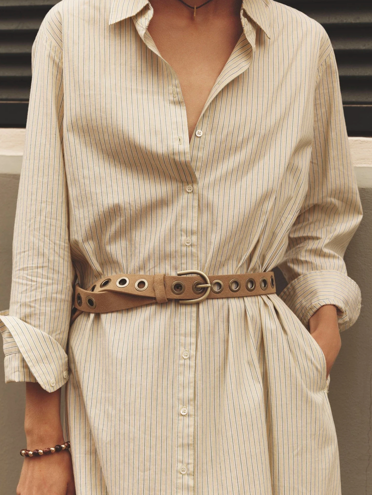 Xirena Boden beige striped cotton dress with rolled sleeves and tan grommet belt, model’s hands in pockets.