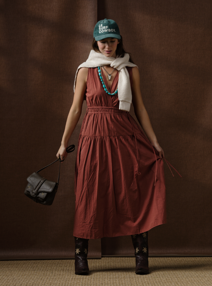 A woman stands before a brown backdrop wearing the Xirena Meri Dress in rust, dark cowboy boots, a white sweater over her shoulders, a green "LIL COWBOY" hat, turquoise necklace, and holding a small black bag.