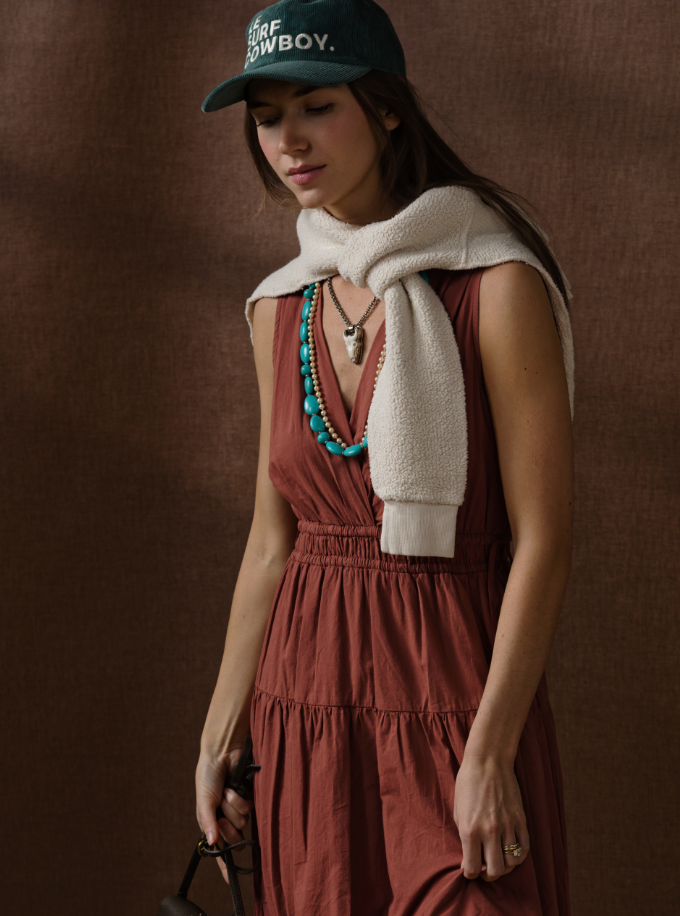 Wearing the Xirena Meri Dress in rust, a woman stands against a brown backdrop with a green cap, a white sweater draped over her shoulders, and layered turquoise and silver necklaces.