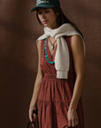 Wearing the Xirena Meri Dress in rust, a woman stands against a brown backdrop with a green cap, a white sweater draped over her shoulders, and layered turquoise and silver necklaces.