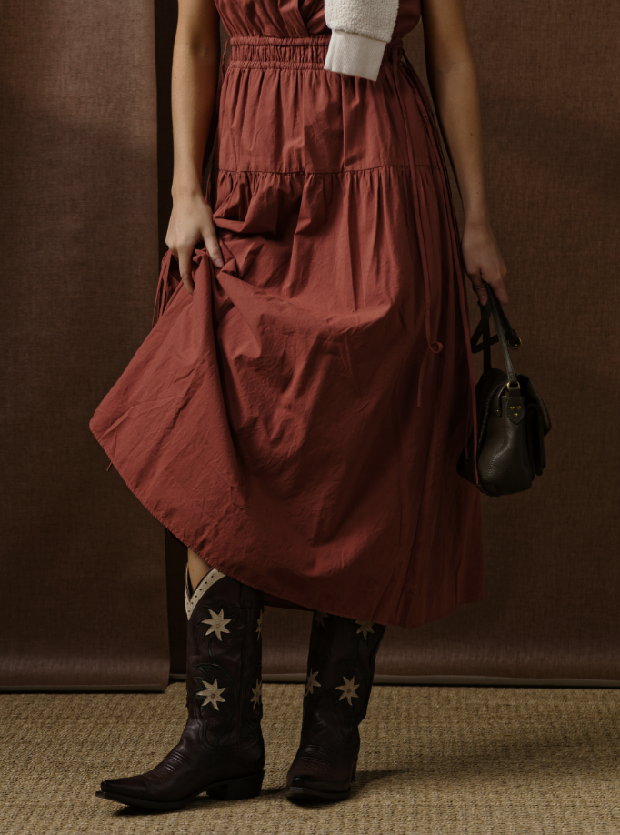 Wearing the Xirena Meri Dress in rust, paired with black floral cowboy boots and a small dark handbag, this look features soft cotton fabric for comfort. The brown background and unseen face keep the focus on the outfit.