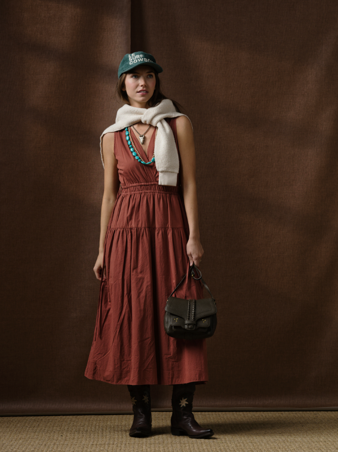 A woman stands before a brown backdrop in the Xirena Meri Dress, paired with cowboy boots, a green cap, turquoise jewelry, a white sweater over her shoulders, and holding a black handbag.