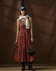 A woman stands before a brown backdrop in the Xirena Meri Dress, paired with cowboy boots, a green cap, turquoise jewelry, a white sweater over her shoulders, and holding a black handbag.