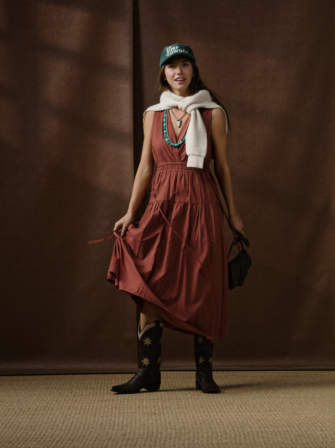 A woman stands indoors wearing the rust-colored Xirena Meri Dress, black cowboy boots with stars, a green hat, turquoise necklaces, a white sweater draped over her shoulders, and carries a small black bag. Brown fabric forms the backdrop.