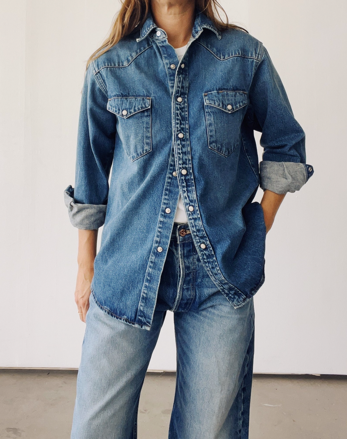 A person wears Hey Gang's The Western Shirt in blue denim over a white top with matching jeans, showing off rolled-up sleeves and a hand-in-pocket pose against a plain white background for a vintage worn look.