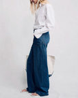 A person with long blonde hair, in a white button-up shirt and Denimist DECK PANT SHANE INDIGO wide-leg jeans, stands in profile against a plain light background, holding a large light tote bag and wearing sandals.