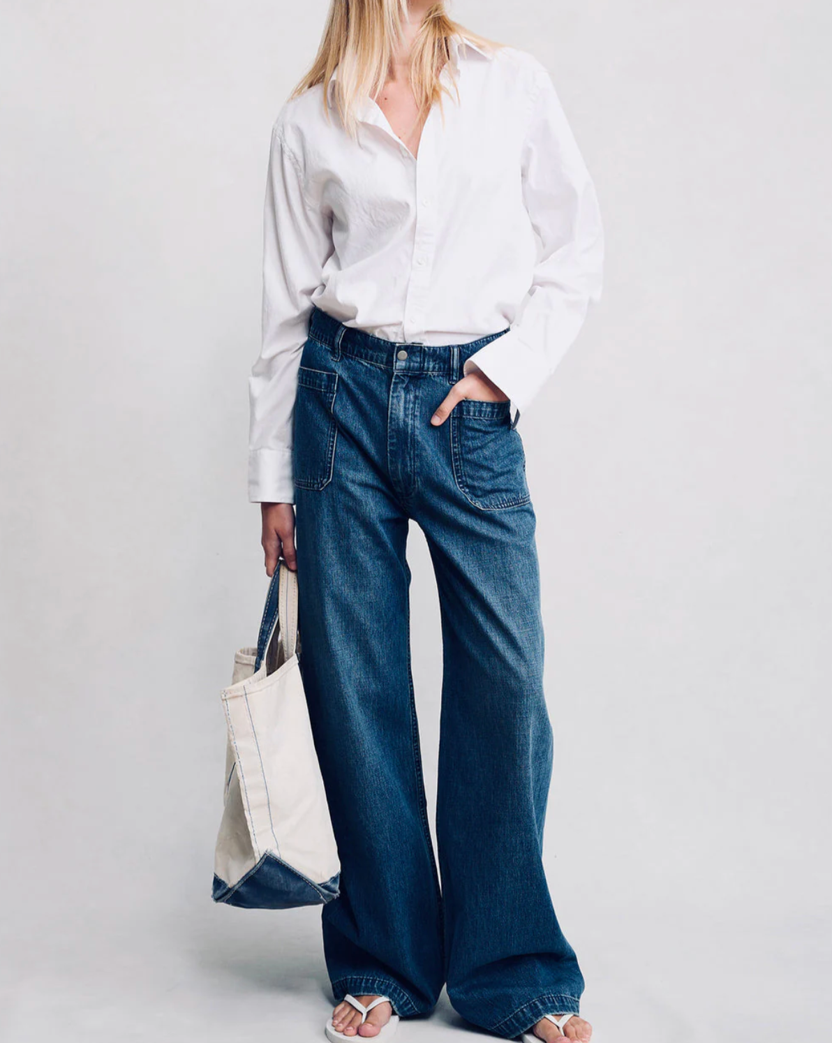 A person in a white button-up shirt, Denimist DECK PANT SHANE INDIGO jeans, and sandals stands holding a beige tote with blue accents against a plain light background. Their face is partially out of frame.