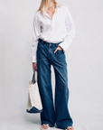 A person in a white button-up shirt, Denimist DECK PANT SHANE INDIGO jeans, and sandals stands holding a beige tote with blue accents against a plain light background. Their face is partially out of frame.