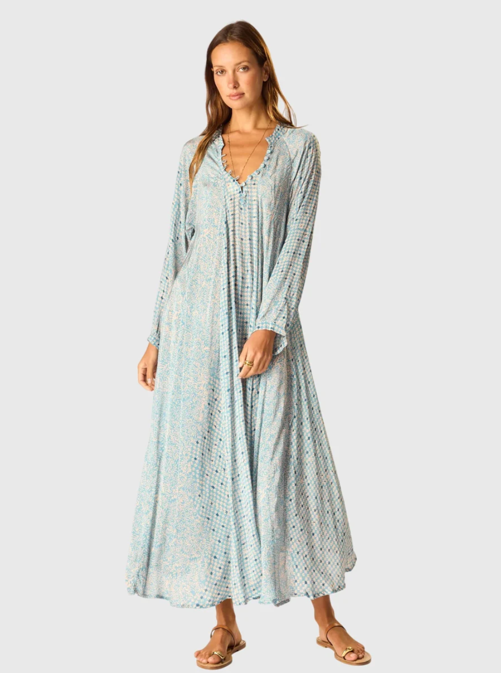 A woman stands barefoot against a plain light gray background, modeling the natalie martin Fiore Maxi—a long sleeve, flowing A-line dress in a light blue pattern with a v-neckline.